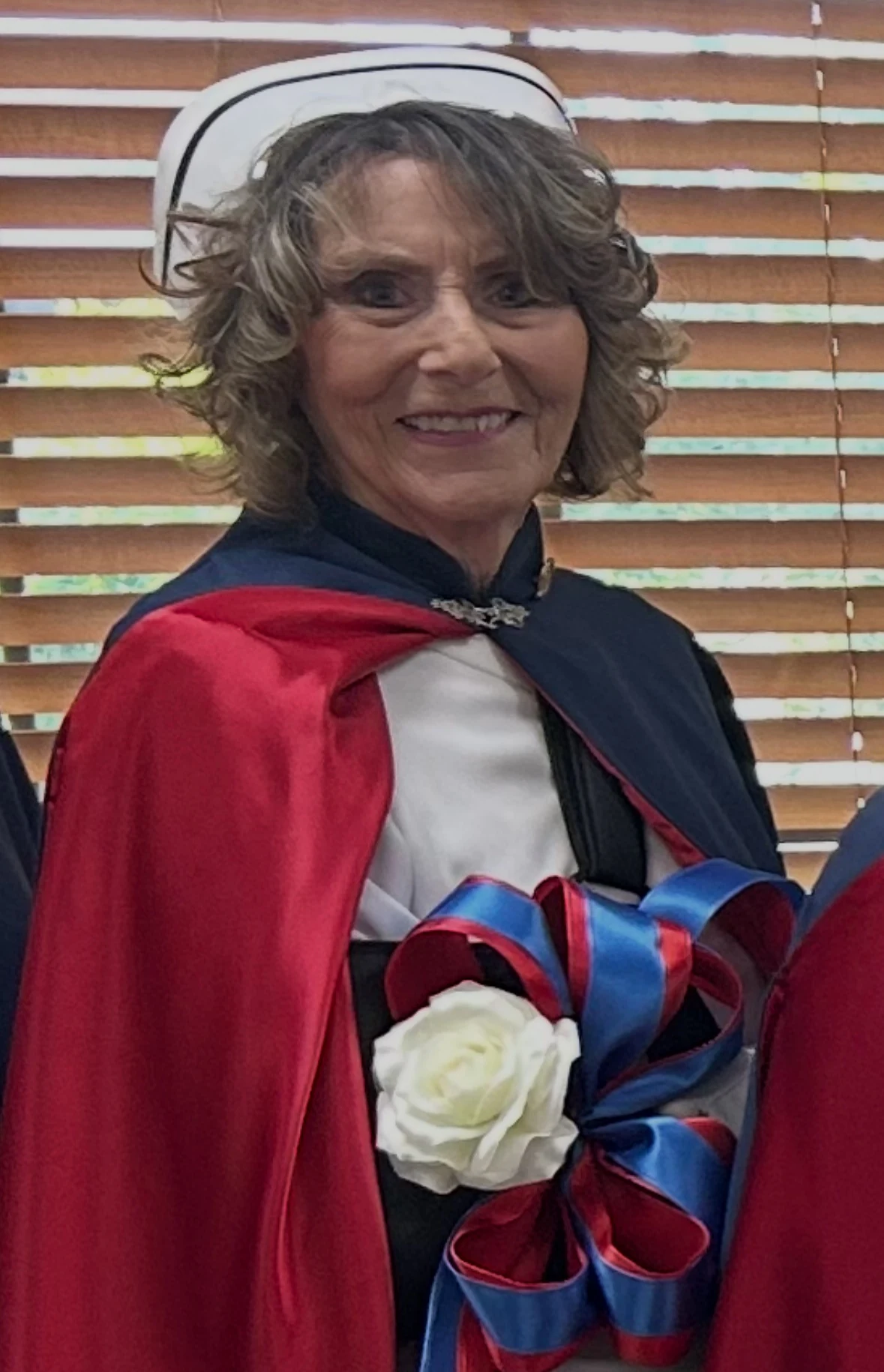 Vicky Lowe in the chapter's white cap and red cape, beside a white rose tied with one of the red and blue bows she makes